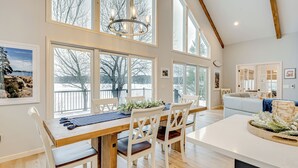 Un comedor espacioso con una mesa de madera y sillas blancas, amplias ventanas que ofrecen vistas al exterior, y una zona de cocina moderna con encimera blanca y una decoración central.