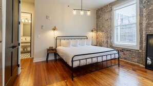 Un dormitorio con estructura de cama metálica, un ventanal con persianas, suelo de madera y pared de ladrillo.