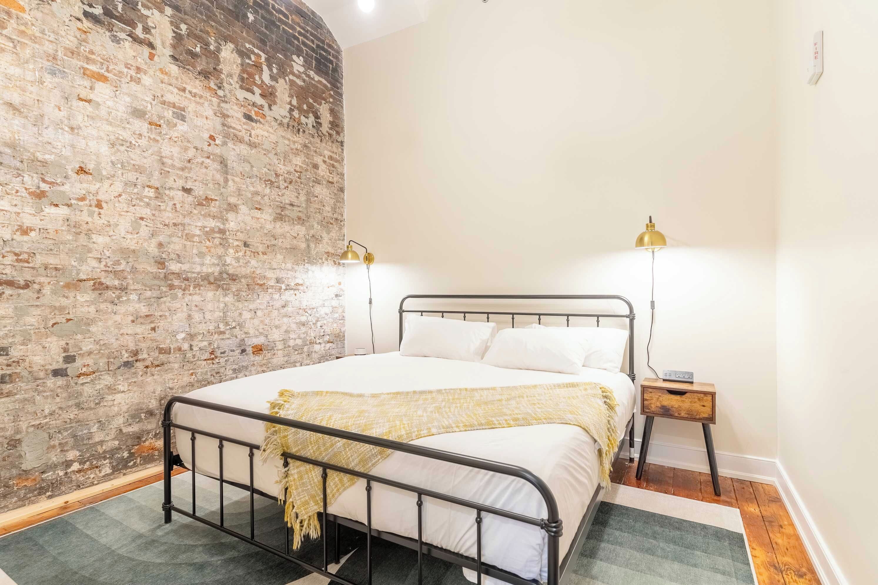 A bedroom with a brick wall, a metal bed frame, two bedside lamps, a bedside table, and a rug.