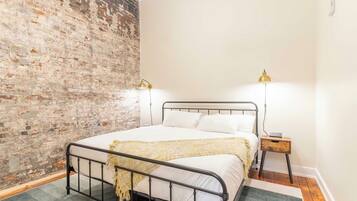 A bedroom with a brick wall, a metal bed frame, two bedside lamps, a bedside table, and a rug.