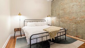 A bedroom with a metal bed frame, white bedding, a brick wall, and two bedside lamps. - 2 Bedroom Suite (Frankfort)