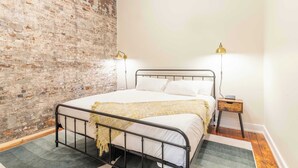 A bedroom with a brick wall, a metal bed frame, two bedside lamps, a bedside table, and a rug. - 2 Bedroom Suite (Frankfort)