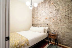 A bedroom with a metal bed frame, white bedding, a brick wall, a small wooden nightstand, and a hanging light fixture. - 2 Bedroom Suite (Frankfort)