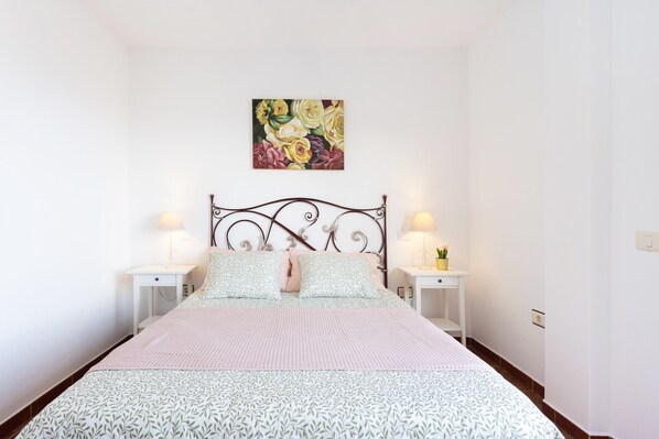 Un dormitorio con una cama, dos mesitas de noche, un cuadro colgado en la pared y una mesita con una lámpara.