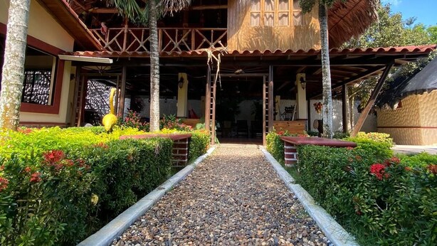 A traditional wooden building with a thatched roof, surrounded by lush greenery and palm trees.