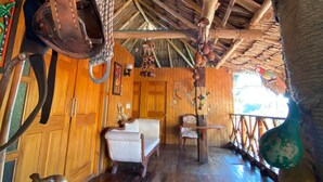 A rustic room with wooden walls and a thatched roof. There are leather bags hanging from the ceiling and a wooden chair in the foreground.