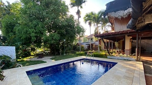 A swimming pool with a tiled deck, surrounded by lush greenery and a thatched-roof structure.