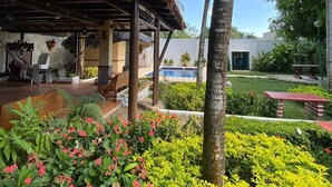 A covered outdoor area with a pool, palm trees, and a variety of plants and flowers.