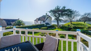 Ein Balkon mit weißen Geländern, ein Korbstuhl mit Kissen und Blick auf ein Haus mit rotem Dach und Schornstein.