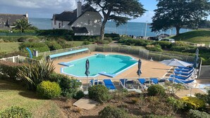 Ein Swimmingpool mit blauen und weißen Liegestühlen, umgeben von einem weißen Zaun, mit Blick auf das Meer und Häuser im Hintergrund.