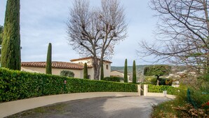 Une allée sinueuse mène à un bâtiment dont le toit est recouvert de tuiles, et qui est entouré de haies soigneusement taillées et de grands arbres.