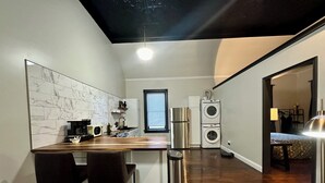 A compact kitchen with a marble backsplash, a wooden dining table with chairs, a refrigerator, a washing machine, and a window with blinds.