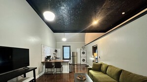 A compact living space with a flat-screen TV, a sofa, a coffee table, and a kitchen area with a refrigerator, washing machine, and a window with blinds.