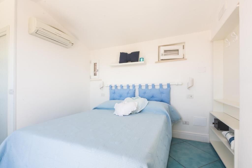 A bedroom with a bed, a white air conditioning unit, a small shelf with a book and a candle, and a white wardrobe.