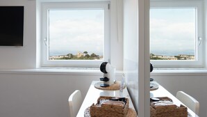 A dining area with a table set for two, two woven baskets, a coffee maker, and a view of the sea and buildings through large windows. - BEAUTY HOME SORRENTO (Sorrento)