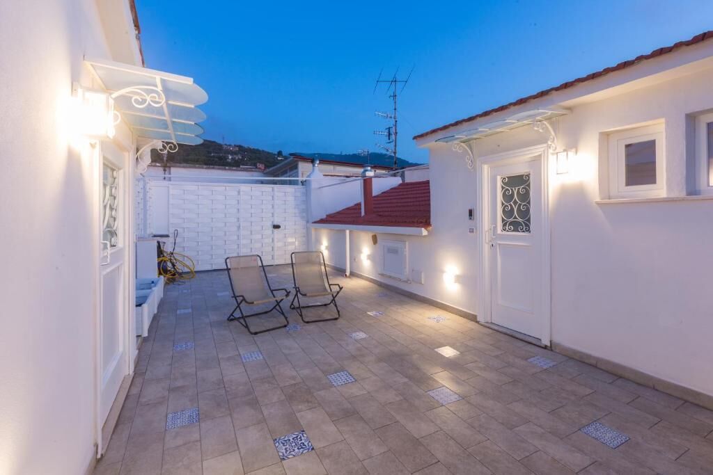 A covered outdoor patio with two lounge chairs, a white wall with a window, and a red roof.