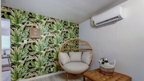 A room with a tropical leaf-pattern wallpaper, a wicker chair with cushions, a wooden bench, and a wall-mounted air conditioner. - The Oakley House (Salt Lake City)