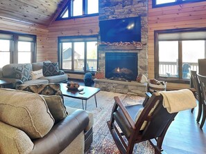 A cozy living room with a stone fireplace, wooden walls, and a large window overlooking a deck.