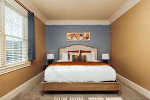 Une chambre à coucher comprenant un lit, deux tables de chevet, une fenêtre avec des stores et une œuvre d’art accrochée au mur.