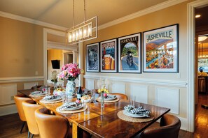 Un coin repas avec une table en bois dressée pour six personnes, entourée de chaises en cuir. Des affiches encadrées ornent le mur, un lustre est suspendu au-dessus de la table et un vase rempli de fleurs est posé sur la table.