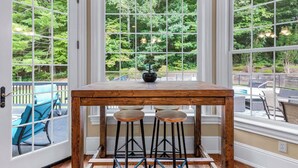 A corner room with a wooden table and two stools, large windows with a view of greenery, and a door with a view of outdoor seating. - 8BR Resort Estate with Pool and Theater (Fletcher)