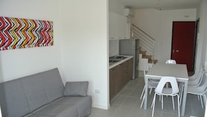 A modern living space with a grey sofa, a dining table with chairs, a kitchen area with a stove and cabinets, and a staircase leading to another level. - Lucia D in Bibione With 3 Bedrooms and 2 Bathrooms (San Michele al Tagliamento)