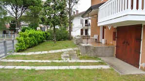 A residential building with a stone pathway leading to a wooden door, surrounded by greenery and a small patio area. - Livia in Bibione With 2 Bedrooms and 2 Bathrooms (San Michele al Tagliamento)