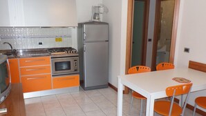 A compact kitchen with a gas stove, oven, refrigerator, and orange cabinets. There's a dining table with four chairs and a colorful rug. - Felice CPT in Bibione (San Michele al Tagliamento)