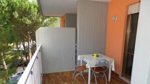 Un balcón con piso de madera, una mesa con sillas y vista a árboles y un coche estacionado.