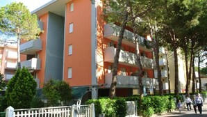 Un edificio residencial de varios pisos con balcones, rodeado de árboles y una cerca blanca.