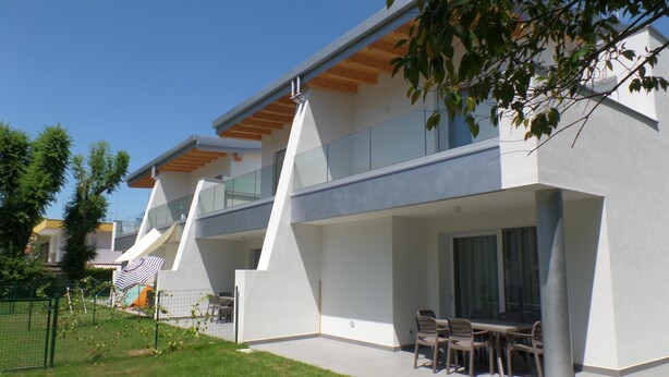 A modern two-story house with a flat roof, large windows, and a patio with outdoor furniture. - Villa Onda D in Bibione (San Michele al Tagliamento)