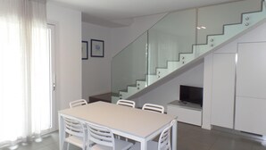 A modern kitchen with white cabinetry, a dining area with a table and chairs, and a glass staircase leading to an upper level. - Villa Onda D in Bibione (San Michele al Tagliamento)