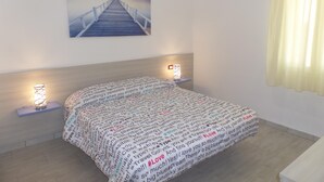 A bedroom with a bed, a headboard, two bedside lamps, a wall-mounted picture, and a window with curtains. - Villa Angi in Bibione (San Michele al Tagliamento)