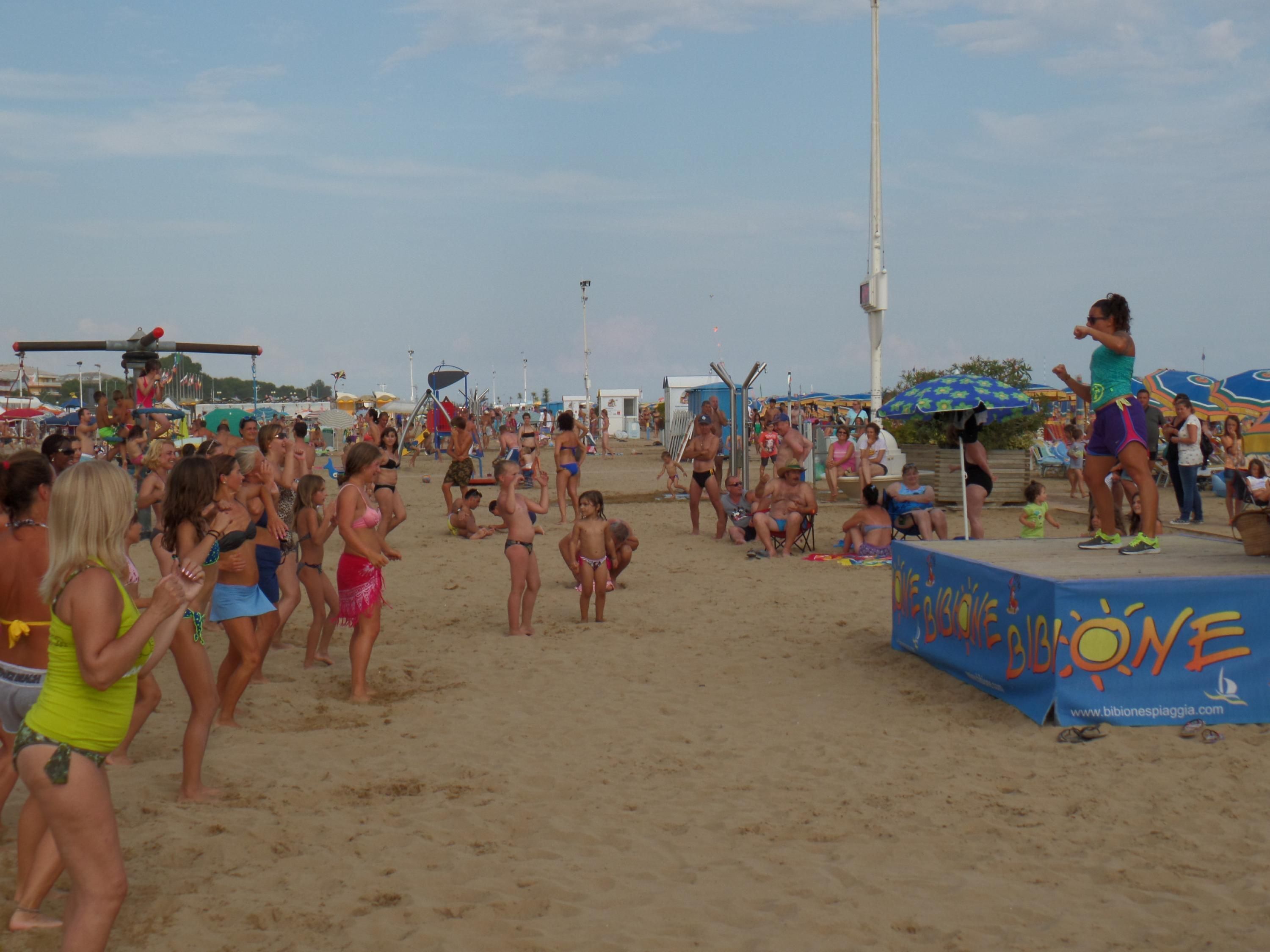 Een druk strand met mensen in zwemkleding, een podium met een spandoek, en strandparasols op de achtergrond.