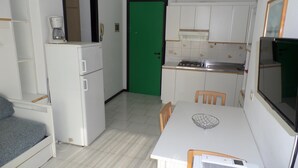 A compact kitchen with a dining area, white cabinets, a green door, and a small refrigerator. - Condominio Luisa in Bibione (San Michele al Tagliamento)