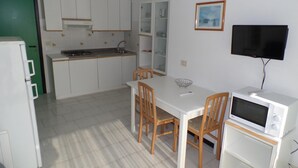 A compact kitchen with white cabinets, a dining table with chairs, a microwave, and a television on the wall. - Condominio Luisa in Bibione (San Michele al Tagliamento)