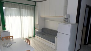 A compact kitchenette with a refrigerator, a microwave, and a coffee maker. There is a small dining table with chairs and a window with curtains. - Condominio Luisa in Bibione (San Michele al Tagliamento)
