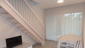 A modern living room with a flat-screen TV on a white cabinet, a striped sofa, a dining table with chairs, and a staircase with white railings. - Melograno C Mans in Bibione (San Michele al Tagliamento)