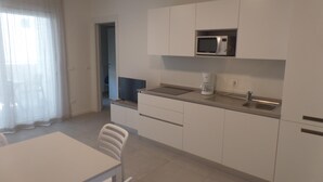 A modern kitchen with white cabinets, a microwave, a coffee maker, and a dining area with a table and chairs. - Melograno A C2 in Bibione (San Michele al Tagliamento)