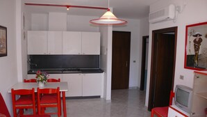 Een moderne keuken met witte kasten en een zwart aanrecht, een eethoek met rode stoelen en een witte tafel, en een televisie op een witte kast.