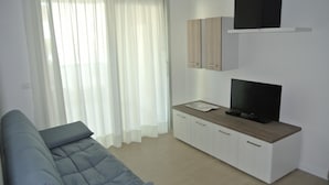 Un salon moderne avec un canapé gris, un téléviseur posé sur un meuble en bois et des rideaux blancs.