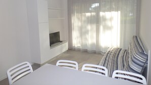 A small, bright room with a white table, chairs, a sofa, a TV on a shelf, and a large window with sheer curtains. - Melograno B B in Bibione (San Michele al Tagliamento)