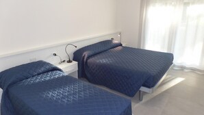 A room with two beds, each with a quilted blue bedspread, a bedside table with a lamp, and a power outlet. - Melograno B B in Bibione (San Michele al Tagliamento)