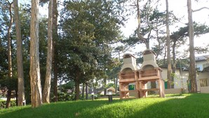 A grassy area with two wooden structures resembling a gazebo or pavilion, surrounded by tall trees and a residential building in the background. - Melograno B B in Bibione (San Michele al Tagliamento)