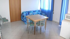 A room with a wooden table and chairs, a blue sofa, a wooden door, and a white refrigerator. - Horizon 16 in Bibione (San Michele al Tagliamento)