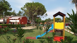 A playground with a slide and climbing structures, surrounded by well-maintained grass and trees. - Azzurro 346 in Bibione (San Michele al Tagliamento)