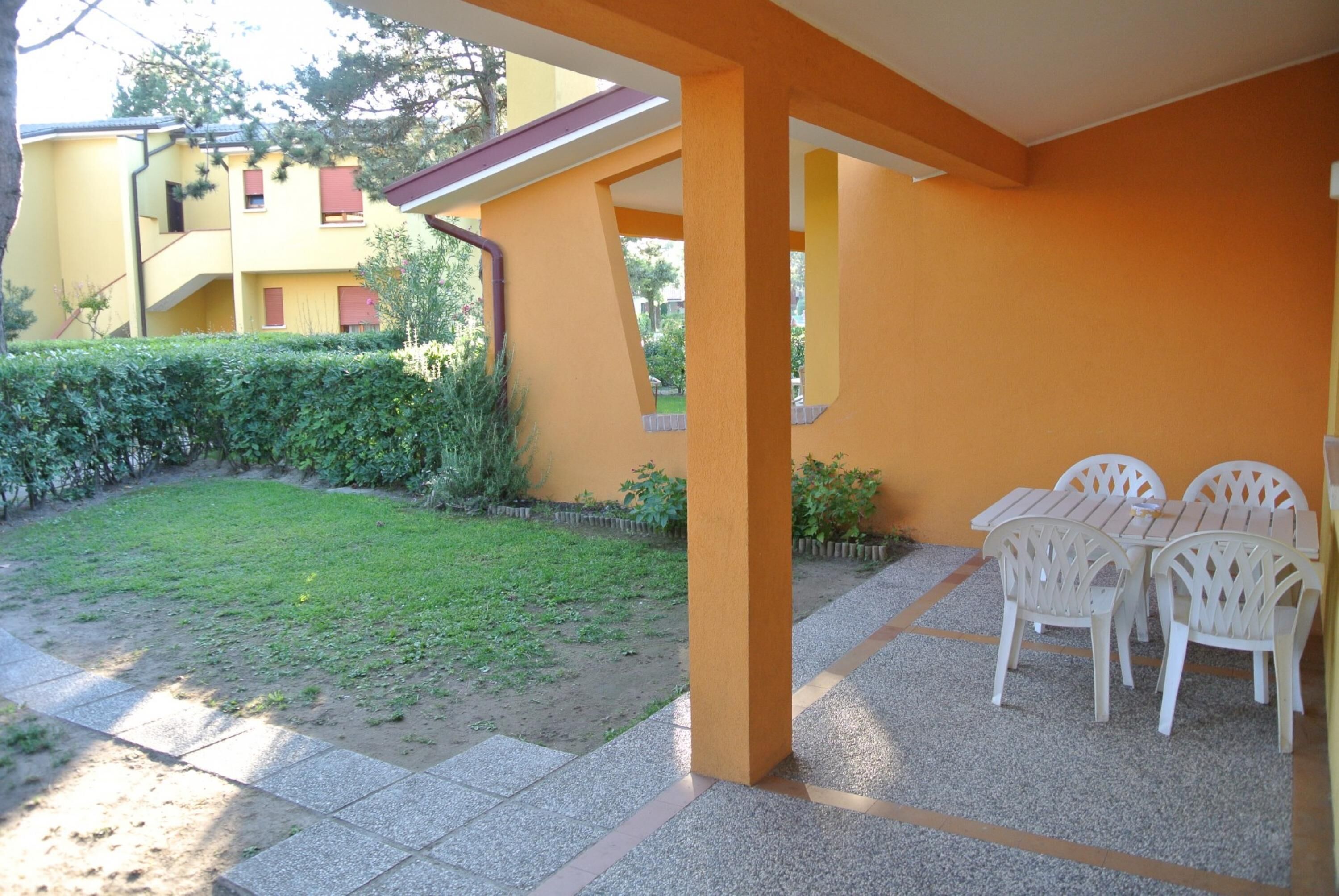 An outdoor patio area with a covered seating area, a grassy lawn, and a yellow building in the background.