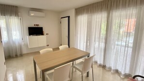 Ein moderner Essbereich mit Holztisch und weißen Stühlen, einem wandmontierten Flachbildfernseher und einem großen Fenster mit durchsichtigen Vorhängen.