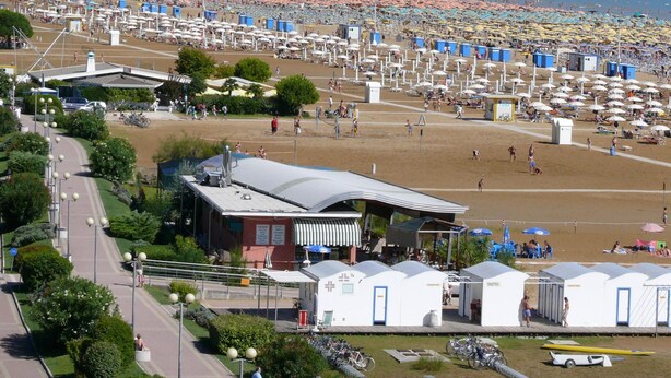 Ein Strand mit vielen Sonnenschirmen und Badegästen. Es gibt mobile Toiletten und ein Gebäude mit dem Schild 'TOILETTES'.