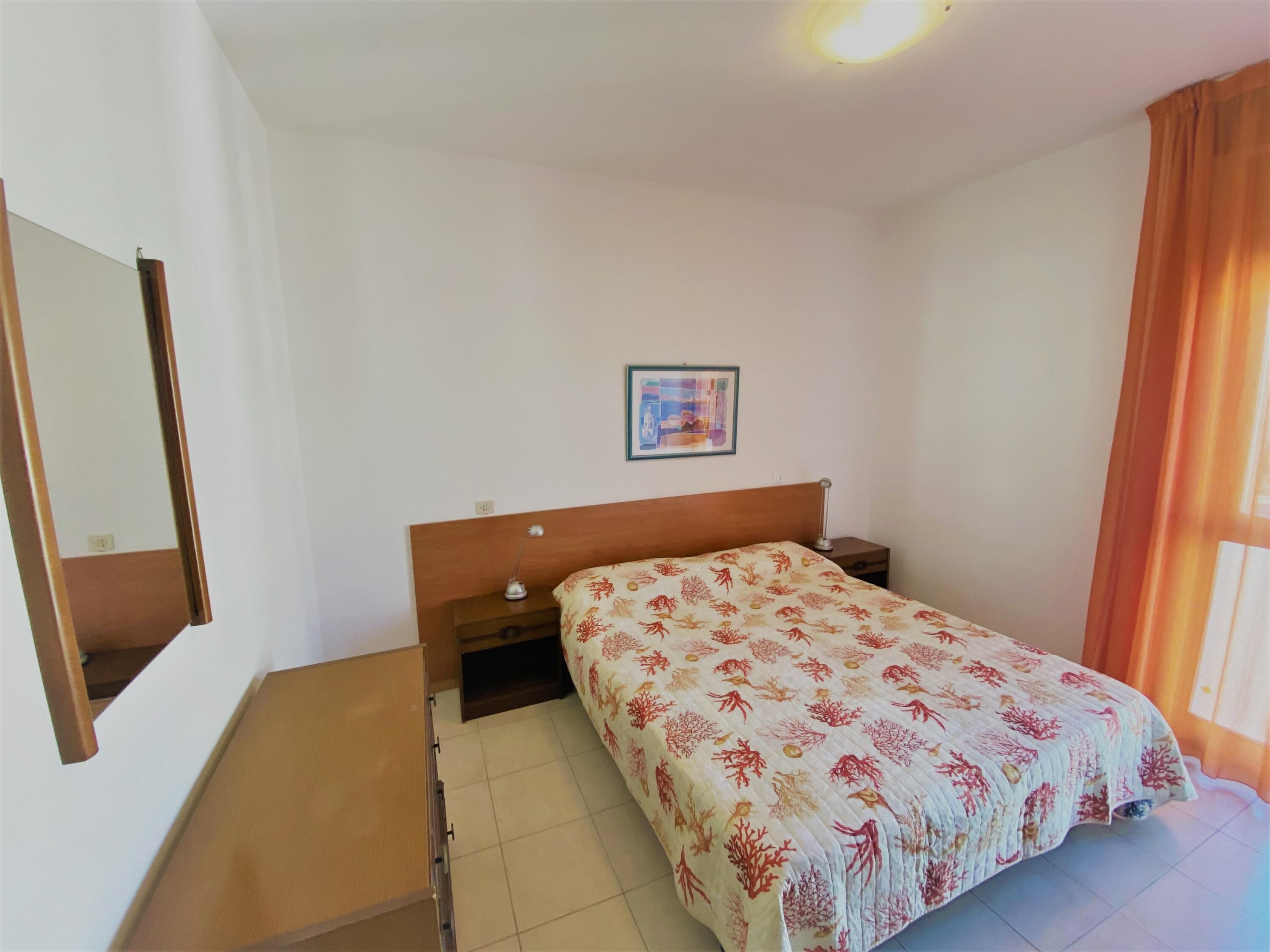 Ein Schlafzimmer mit Bett, Nachttisch, Lampe, einem Wandbild und einem Spiegel.